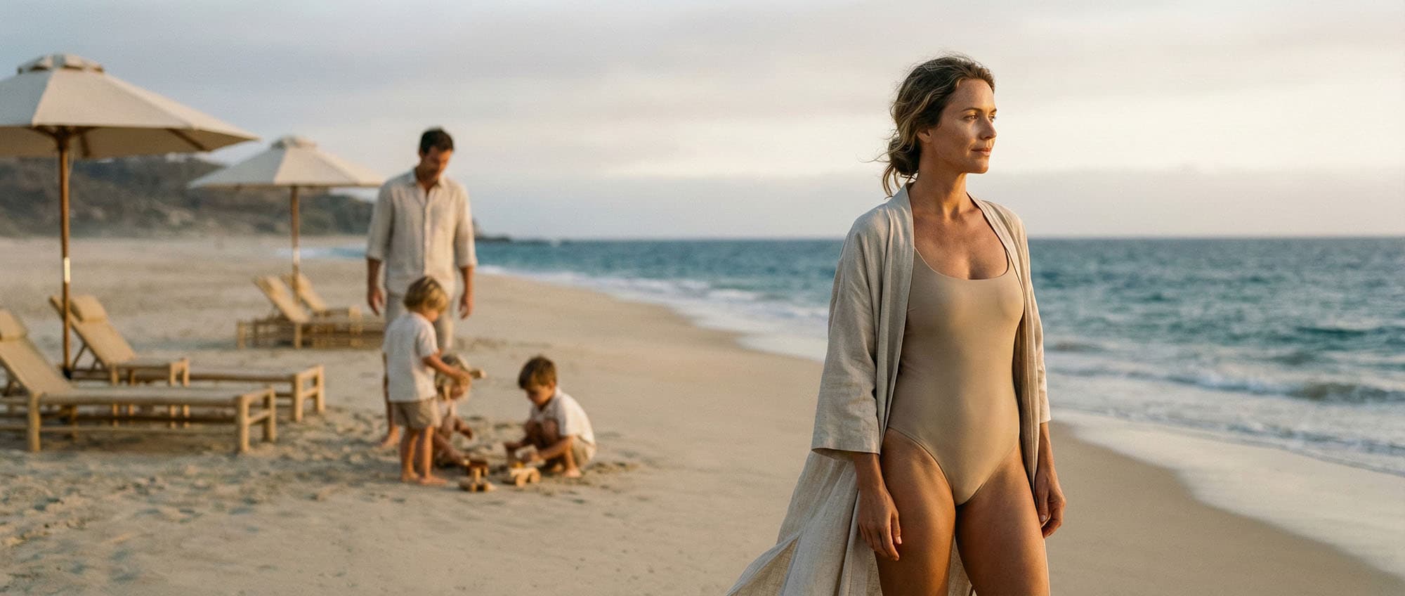 A woman in a beige swimsuit and cover-up stands on a sandy beach, while a man and two children play near lounge chairs and umbrellas by the ocean.
