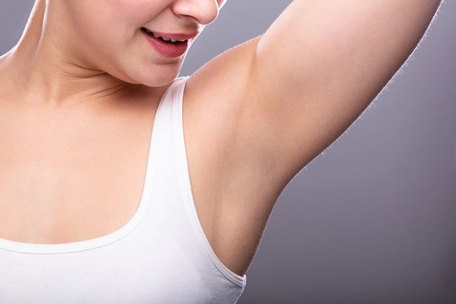 A woman in a sleeveless black top lifts one arm and touches her opposite shoulder, subtly highlighting her smooth underarm—an inspiring look for those considering Armpit Fat Removal in NYC. She poses with pursed lips against a white background.