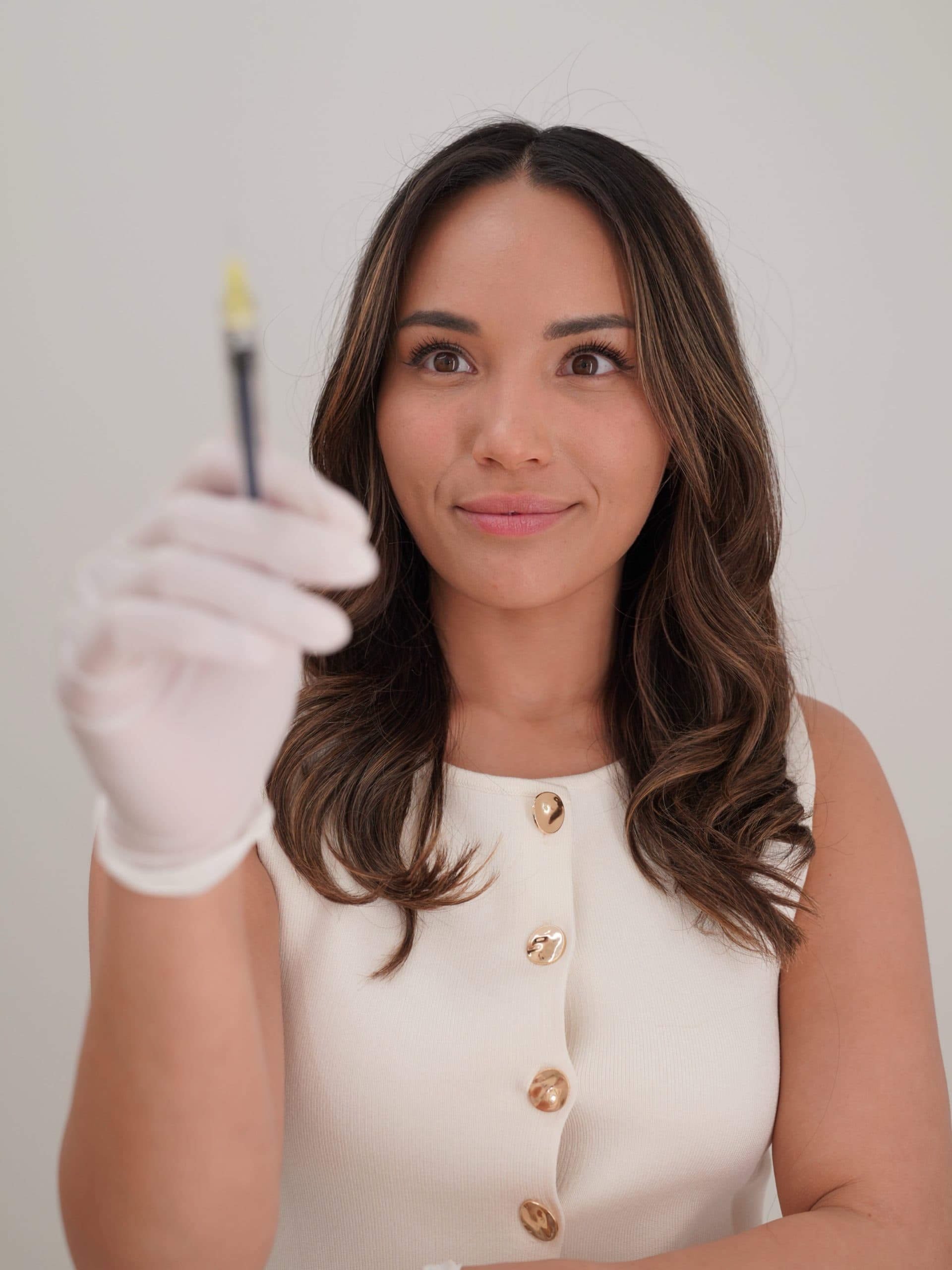 Woman wearing white gloves holds up a syringe, looking at it intently. She is dressed in a sleeveless white top with gold buttons.