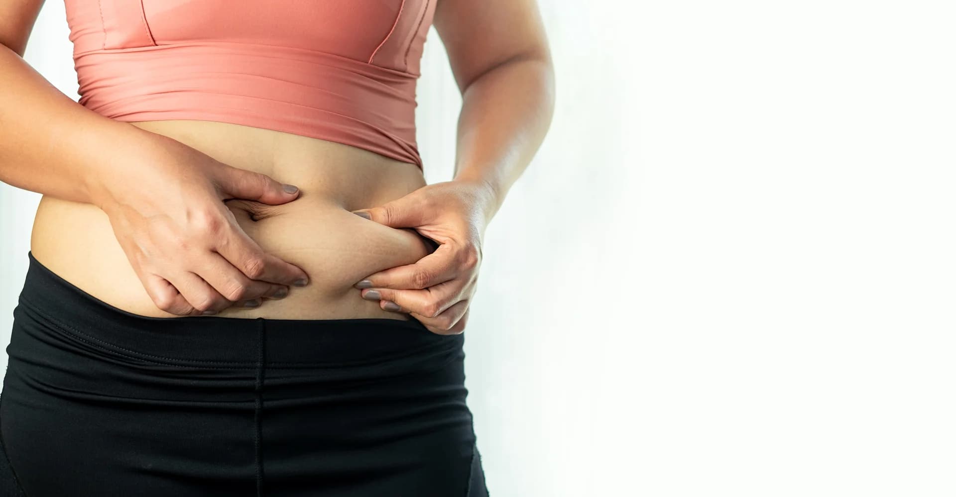 Woman pinching belly fat, wearing a pink top and black pants, post Lipo 360 surgery, against a white background.