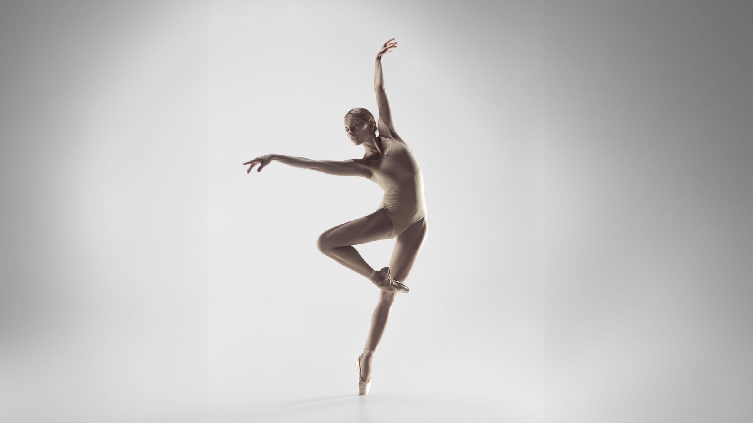 A ballerina in a beige leotard balances on one pointe, with one leg raised and arms gracefully extended against a plain, light background.