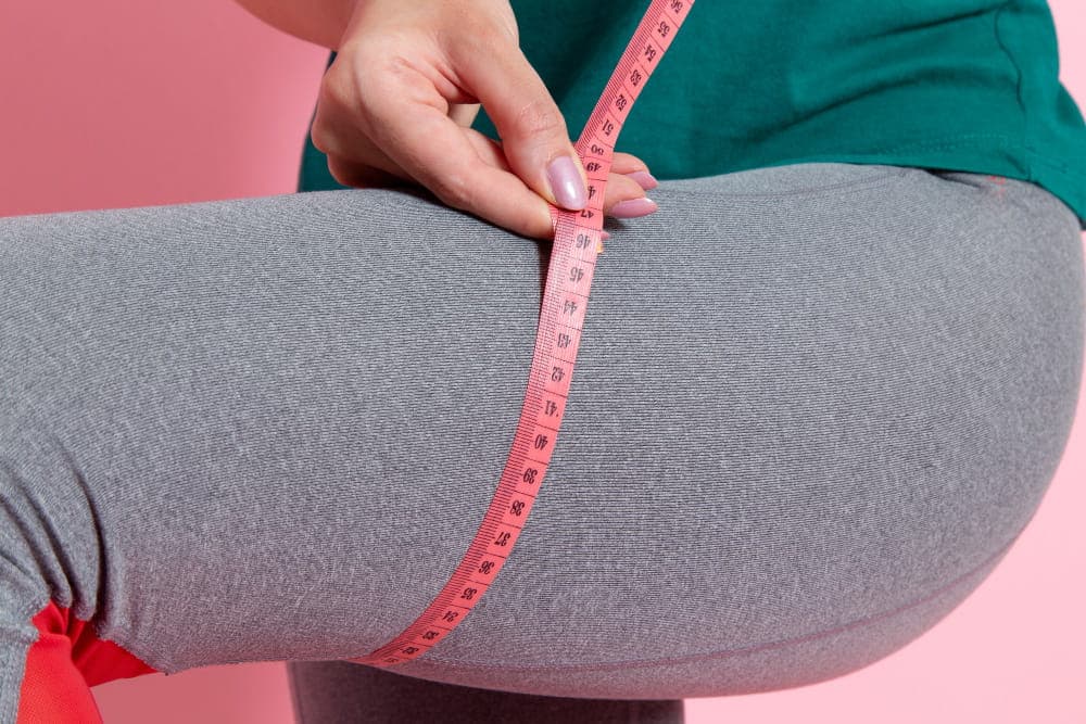 A person in gray leggings measures their thigh circumference with a pink measuring tape against a pink background, highlighting differences that may indicate conditions like obesity or lipedema.