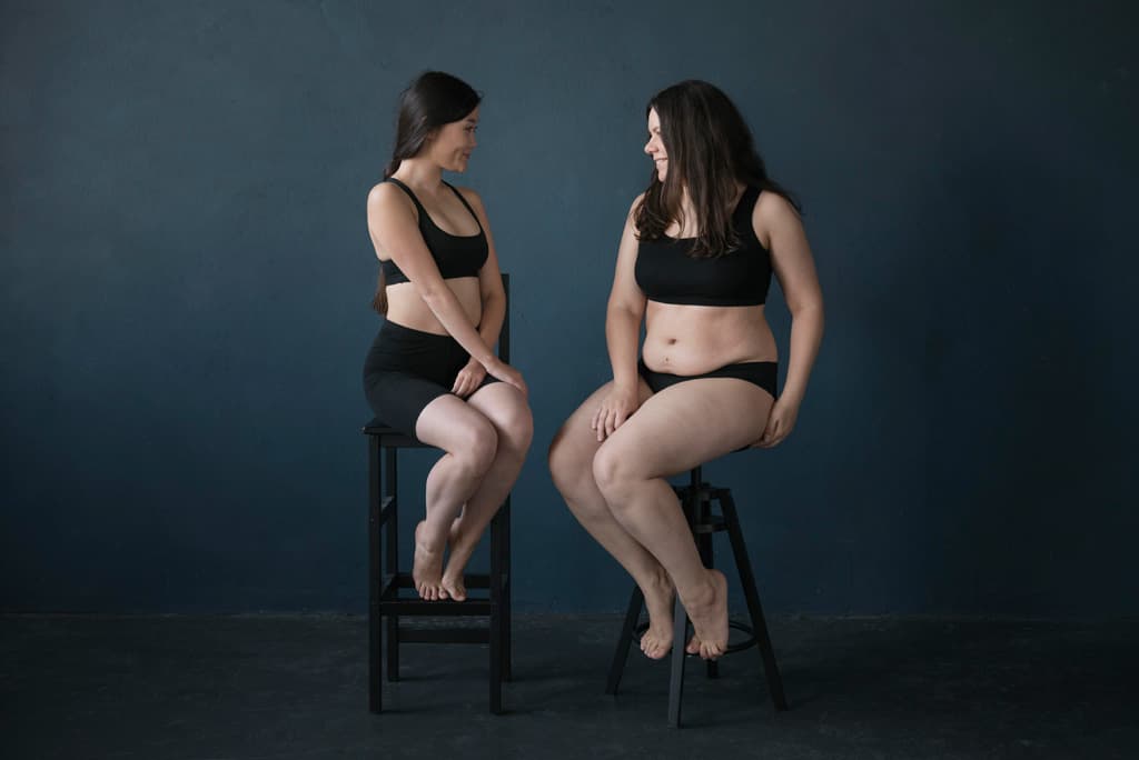 Two women in black underwear sit on stools facing each other, smiling, against a plain dark blue background—celebrating their differences and raising awareness for Lipedema and Lymphedema.