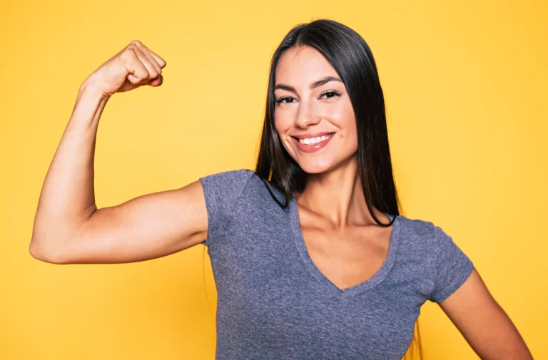 A young woman flexing her toned arms on a yellow background.