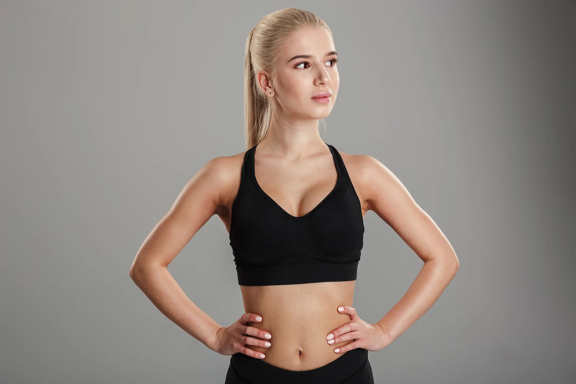 A young woman in a black sports bra showcasing precise arm lipo results on a gray background.
