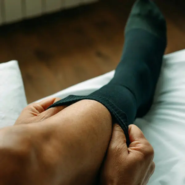 A man is putting calf-length socks on his feet on a bed.