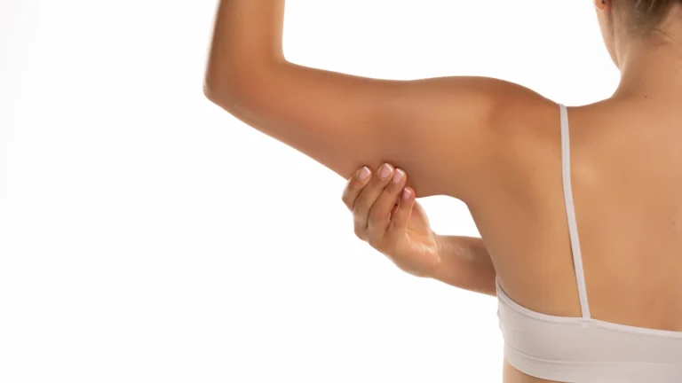 A woman in a white sports bra checking her tricep muscle before treatment for loose skin, isolated on a white background.